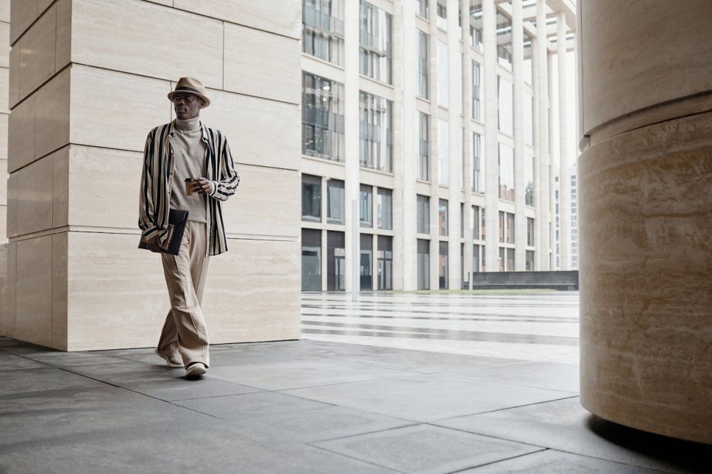 Fashionable man wearing hat and striped jacket walking in a modern cityscape.
